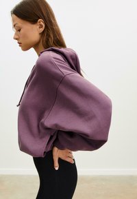 Ljubičasti oversized dukserica s kapuljačom, širokim rukavima i mekanom teksturom. Nosena s crnim tajicama, naglašavajući opuštenu siluetu.