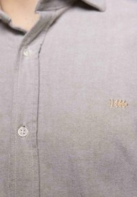 Camisa de botones de color gris claro con una textura suave, que presenta un pequeño detalle bordado en oro y un diseño de cuello clásico.