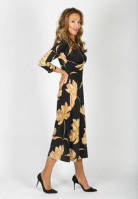 Musta mekko, jossa on kultaiset kukkakuviot, pitkät hihat, polveen ulottuva, istuva yläosa, valuväri, yhdistetty mustien korkokenkien kanssa.