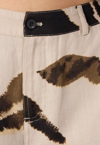 Beige farkkukankaat sisältävät mustan nappikiinnityksen ja abstrakteja ruskeita kuvioita. Saumojen näkyvä ommel tuo yksityiskohtia kankaaseen.