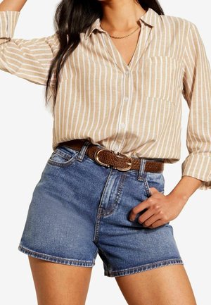 Beżowo-biała koszula w paski z guzikami, włożona do wysokich, niebieskich szortów jeansowych z brązowym teksturowanym paskiem, noszona przez kobietę.