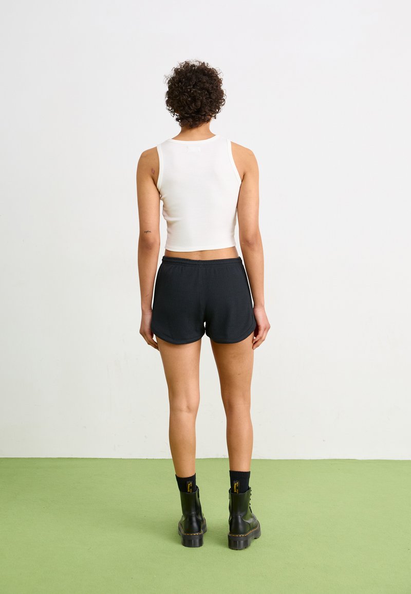 Weißes, kurzes Tanktop, schwarze, gerippte Shorts, schwarze Kampfstiefel. Das Model ist von hinten auf einem hellgrünen Boden zu sehen.