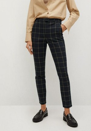 Slim-fit zwarte broek met beige ruitpatroon, gedragen met een beige overhemd en zwarte loafers, hand in de zak, staand tegen een witte achtergrond.