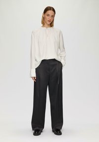 Weiße Bluse mit langen Ärmeln, gerafftem Halsauschnitt und Schlüssellochdetails, kombiniert mit locker sitzenden schwarzen Hosen und Plateauschuhen.