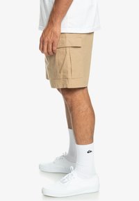 Bermuda cargo beige avec un motif texturé, une poche latérale et une coupe droite. Assorti à des baskets blanches et des chaussettes blanches jusqu'aux genoux.