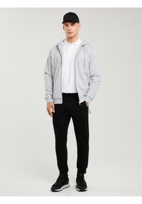 Mees, kes kannab musta nokkmütsi, valget polo, helehallil lukuga kapuutsi, mustad dressipükse ja musti tossusid, seisab tavalise tausta ees.