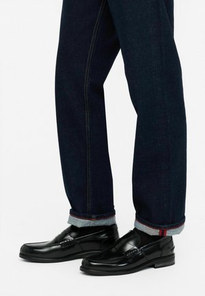 Mocasines de cuero negro con un acabado pulido, combinados con jeans de mezclilla oscura con dobladillo y costuras rojas en contraste.