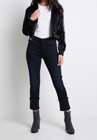 Black glossy bomber jacket over a white T-shirt, paired with dark blue jeans featuring frayed hems, and gray high-heeled ankle boots.