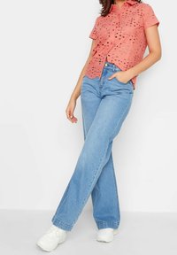 Kobieta ubrana w koralową bluzkę z krótkim rękawem z otworkami, jasnoniebieskie dżinsy z wysokim stanem, białe sneakersy, z jedną ręką w kieszeni i skrzyżowanymi nogami.