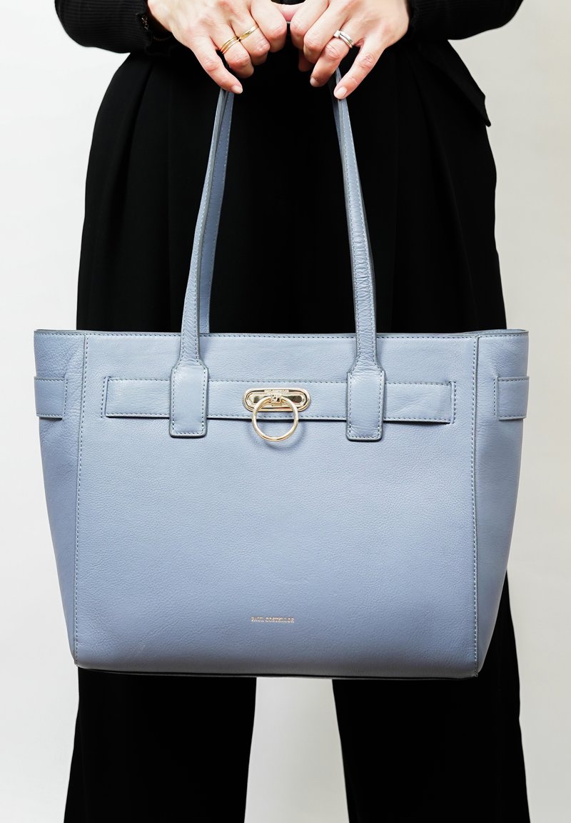 Lichtblauwe leren tote bag met dubbele handgrepen, voorzien van een gouden cirkelvormige hardware accent en een zachte textuur.