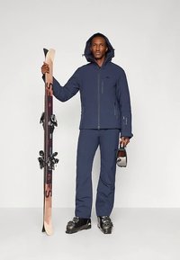 Άνδρας ντυμένος με σκούρο μπλε στολή σκι και μπότες, κρατώντας τα σκι στο ένα χέρι και τις γυαλιά σκι στο άλλο, στέκεται μπροστά από απλό φόντο.