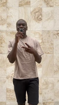 Šviesiai beige marškinėliai su lygiu paviršiumi, derinami su juodais šortais. Figūra laiko mikrofoną ir rodo taikos ženklą.