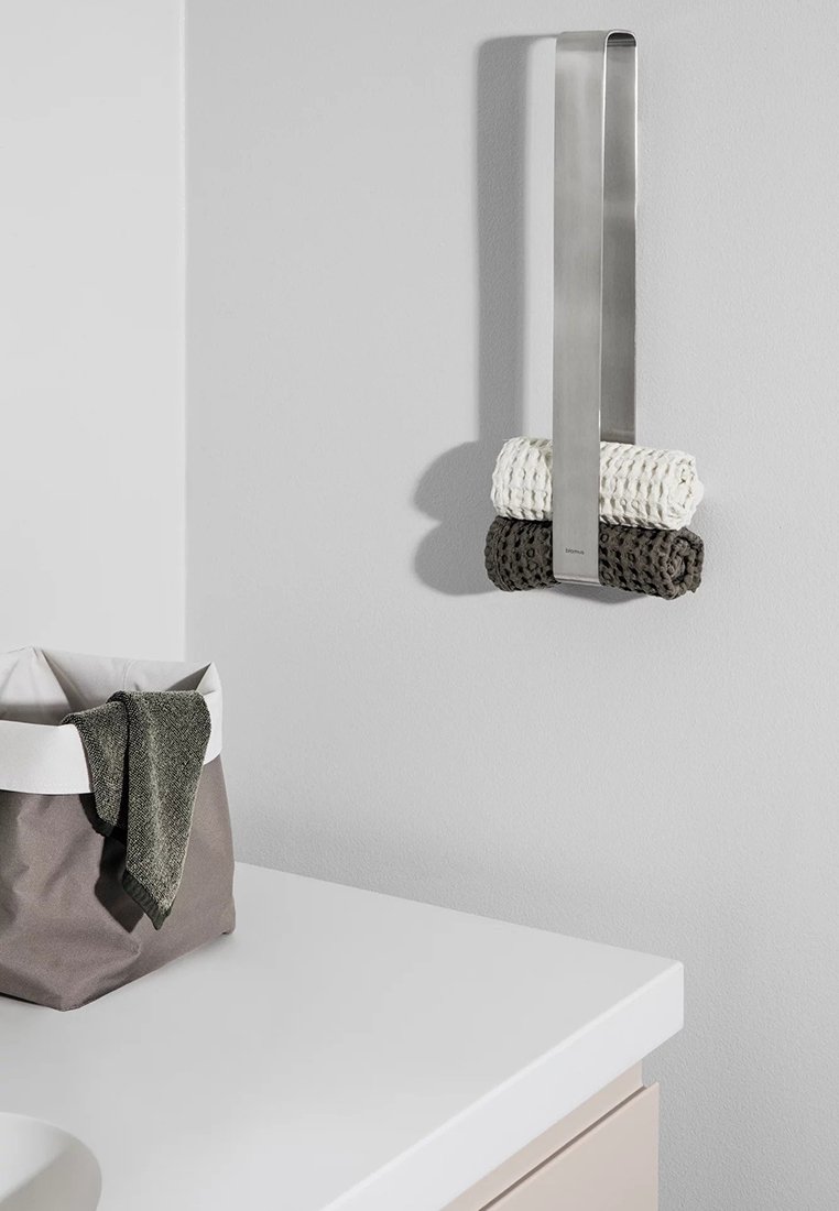 Edelstahl-Handtuchhalter an einer Wand montiert, mit gerollten Handtüchern in Weiß und Dunkelgrau. In der Nähe befindet sich ein Aufbewahrungskorb aus strukturiertem Stoff.