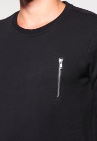 Sudadera negra de mezcla de algodón con cuello redondo, que presenta un bolsillo con cremallera plateada en el pecho izquierdo y tejido texturizado.