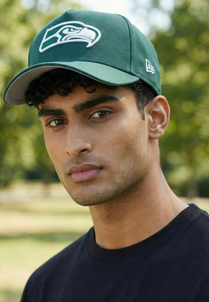 Junger Mann mit lockigem Haar, der eine grüne Seattle Seahawks Kappe und ein schwarzes Shirt trägt, draußen mit unscharfem grünem Baumhintergrund.