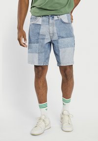 Denim shorts featuring a patchwork design with varying shades of blue, frayed edges, and traditional belt loops. Worn with white sneakers and striped socks.