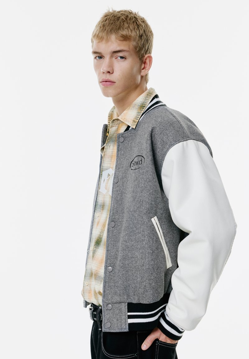 Veste varsity en laine grise et cuir blanc avec des poignets côtelés rayés et un écusson logo. Superposée sur une chemise à carreaux.
