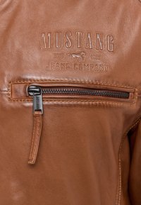Braune Lederjacke mit geprägt "Mustang Jeans Company" Logo, horizontaler Reißverschluss-Tasche mit schwarzem Zubehör und kontrastierenden Nähten.