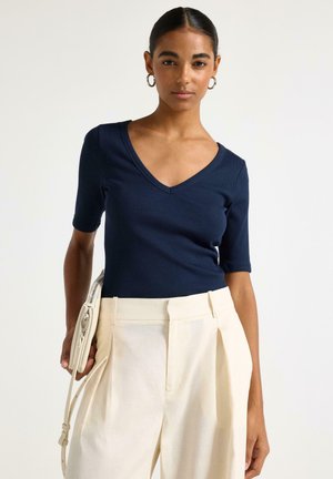 Kvinde iført navyblå V-hals top og højtaljede cremefarvede plisserede bukser, der holder en cremefarvet crossbody-taske med metal lynlås.