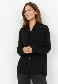 Blusa negra de manga larga con cuello de lazo, con detalles de encaje en los hombros y un corte relajado. Tejido suave y cierre de botones.