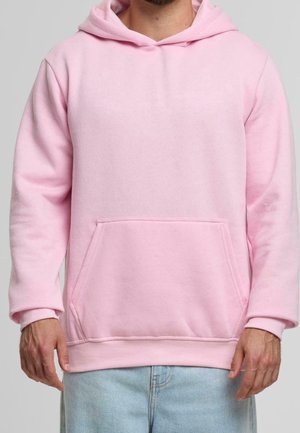 Mann trägt einen hellrosa Kapuzenpullover mit vorne liegender Kängurutasche und hellblaue Jeans vor einem schlichten Hintergrund.