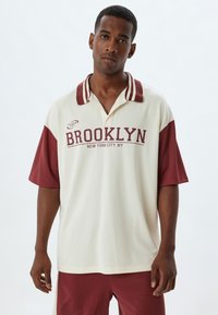 Koszulka polo z krótkim rękawem w kolorze kremowym z bordowymi akcentami i kołnierzykiem. Na piersi znajduje się grafika "BROOKLYN" oraz logo marki sportowej.