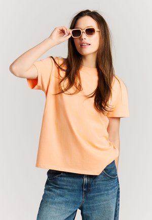 Jeune femme en T-shirt pêche et jean bleu ajustant des lunettes de soleil beiges, debout devant un fond clair uni.