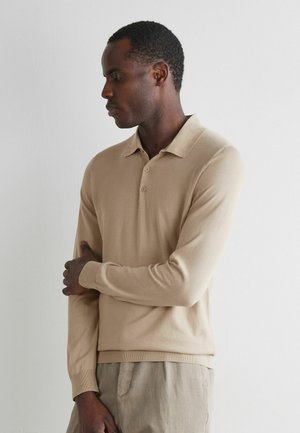 Mann trägt beige langärmliges Polohemd und helle Hose, blickt nach unten mit einem verschränkten Arm und der Hand am gegenüberliegenden Ellbogen.