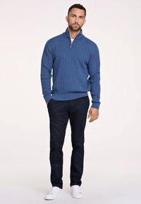 Pull en maille bleu avec un col zippé jusqu'à un quart, associé à un pantalon sombre et des baskets blanches. Texture lisse, coupe décontractée.