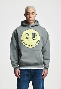 Grå hettegenser med et stort svart og gult sirkulært grafisk design med teksten "2Y f*ck dine dårlige vibber" og en frontlomme.