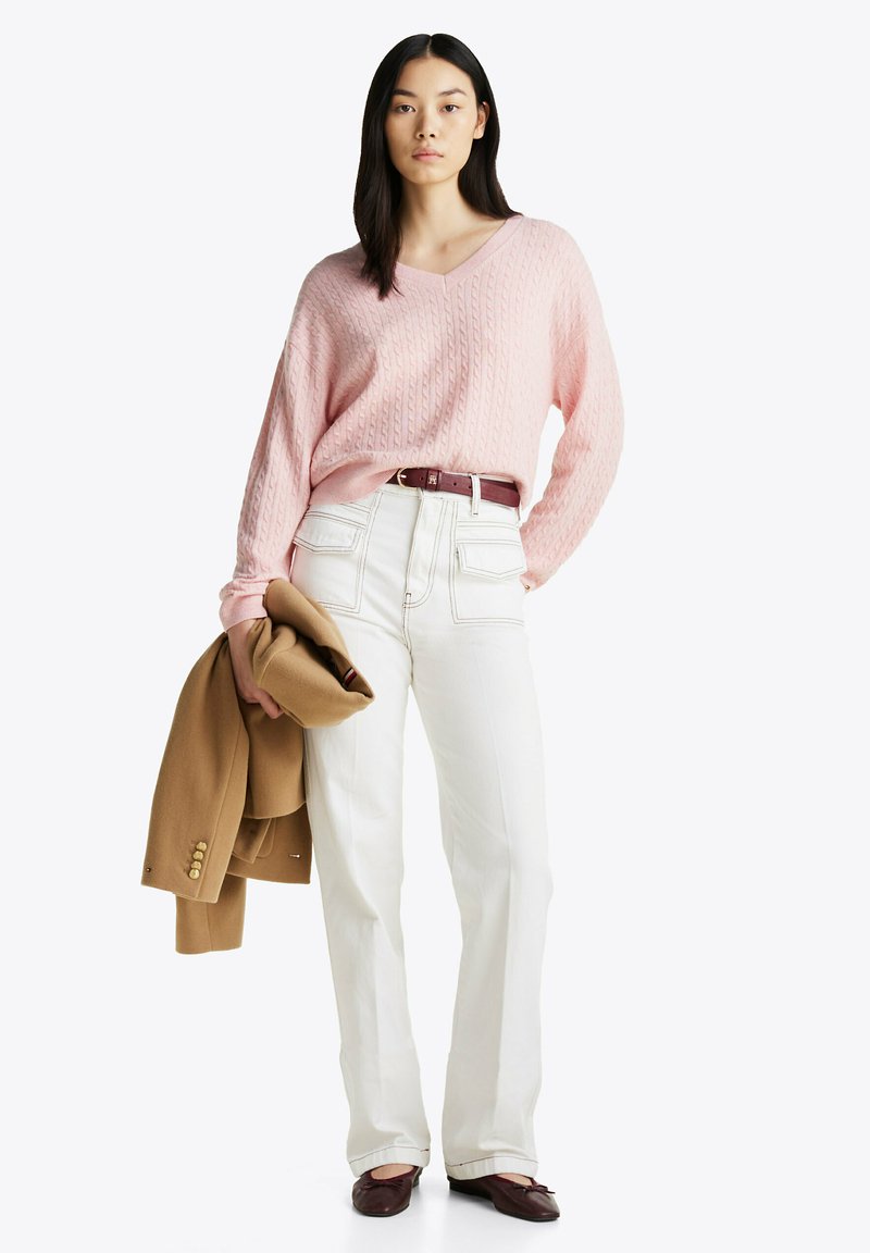 Lichtroze kabelgebreide trui, witte broek met hoge taille, bruine riem en een tan kleurige jas over één arm, gecombineerd met donkere schoenen.