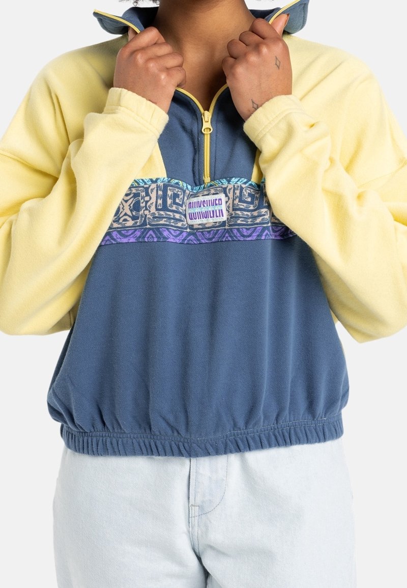 Personne portant une polaire jaune et bleue à fermeture éclair quart, avec un panneau sur la poitrine à motifs et les mains saisissant le col.