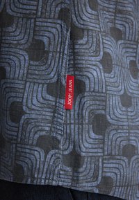Patroon denim stof met onderling verbonden gebogen lijnen in blauwtinten, met een rode "JOOP! JEANS" label gestikt aan de zijnaad.