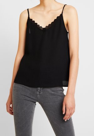 Woman wearing black lace-trimmed spaghetti strap camisole and grey jeans, standing against a plain light background.