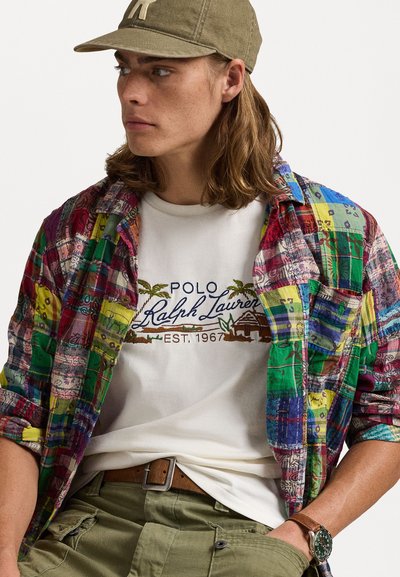 Cămașă din flanel în carouri multicolore peste un tricou alb cu logo brodat, asociată cu pantaloni de culoare verde măsliniu și o curea maro.