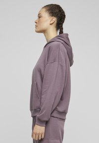 Fioletowa bluza z luźnym krojem, przednią kieszenią, ryflowanymi mankietami i dołem, wykonana z miękkiego materiału. Posiada kaptur ze sznurkiem i minimalistyczny design.