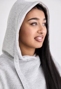 Grå hettegenser med en stor, myk hette, med dobbeltsydd halslinning. Glatt tekstur og snor-detaljer. Modellen smiler mykt.