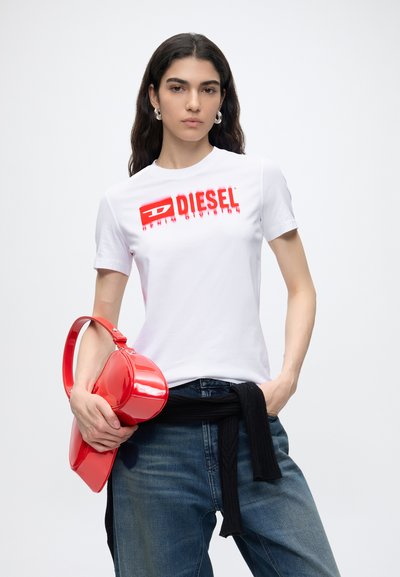 Жена, носеща бяла тениска с лого на Diesel, сини дънки, черен пуловер, завързан на кръста, държащ бляскава червена чанта с една ръка.