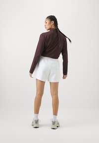 Karamelės spalvos ilgomis rankovėmis marškiniai, derinami su baltais šortais. Marškiniai turi apykaklę; šortai su elastingu juosmeniu. Modelis stovi su nugarą į priekį.