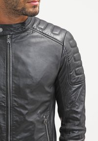 Chaqueta de motocicleta de cuero negro con un diseño estructurado, que presenta acentos acolchados en los hombros, un cierre frontal con cremallera y bolsillos laterales.