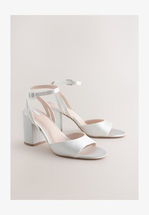 Valged avatud varbaga plokktallaga sandaalid, millel on pahkluu rihmad ja hõbedased pandlad neutraalsel beežil taustal.