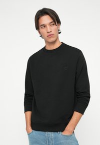 Čierny sweatshirtník s rebrovaným výstrihom a manžetami, s jemne embosovaným logom na hrudi. Hladká textúra, voľný strih a dlhé rukávy.