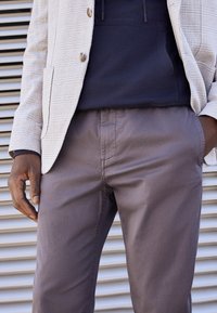 Ljusgrå blazer med en texturerad tyg, bärs över en marinblå hoodie. Grå byxor med en slät yta, hand vilande på höften.