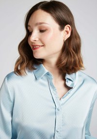 Blouse en satin bleu clair avec un col pointu, présentant un motif à pois texturé subtil et des boutons, créant une finition lisse.