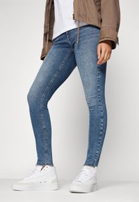 Jeans ajustados en denim azul medio con una textura suave, un desvanecimiento sutil y marcas de desgaste. Combinados con zapatillas blancas de caña alta.