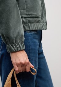 Green corduroy jacket with ribbed texture and elastic cuffs, paired with blue denim jeans. Hand holds a tan leather strap.