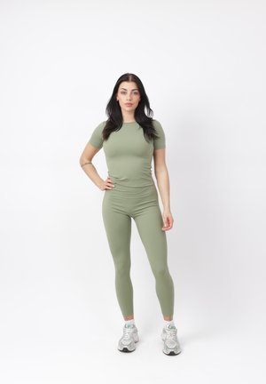 Frau mit langen dunklen Haaren steht frontal, trägt ein eng anliegendes olivgrünes Kurzarmshirt und passende Leggings sowie weiße Turnschuhe.