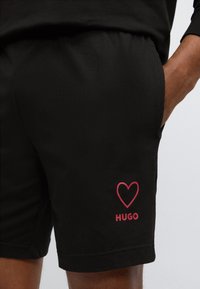 Μαύρα σορτς με ελαστική μέση, πλαϊνές τσέπες και κόκκινη καρδιά με το κείμενο "HUGO" στο αριστερό πόδι.