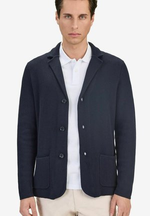 Mies, jolla on päällään laivastonsininen teksturoitu bleiseri valkoisen kauluspaidan ja beigejen housujen kanssa, seisoo yksiväristä valkoista taustaa vasten.