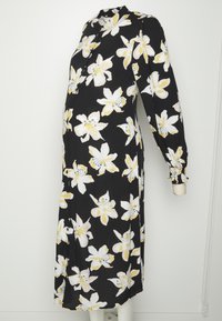 Vestido floral preto com mangas longas, apresentando padrões de lírios brancos e amarelos, fechamento com botões e punhos franzidos, projetado para vestuário de maternidade.